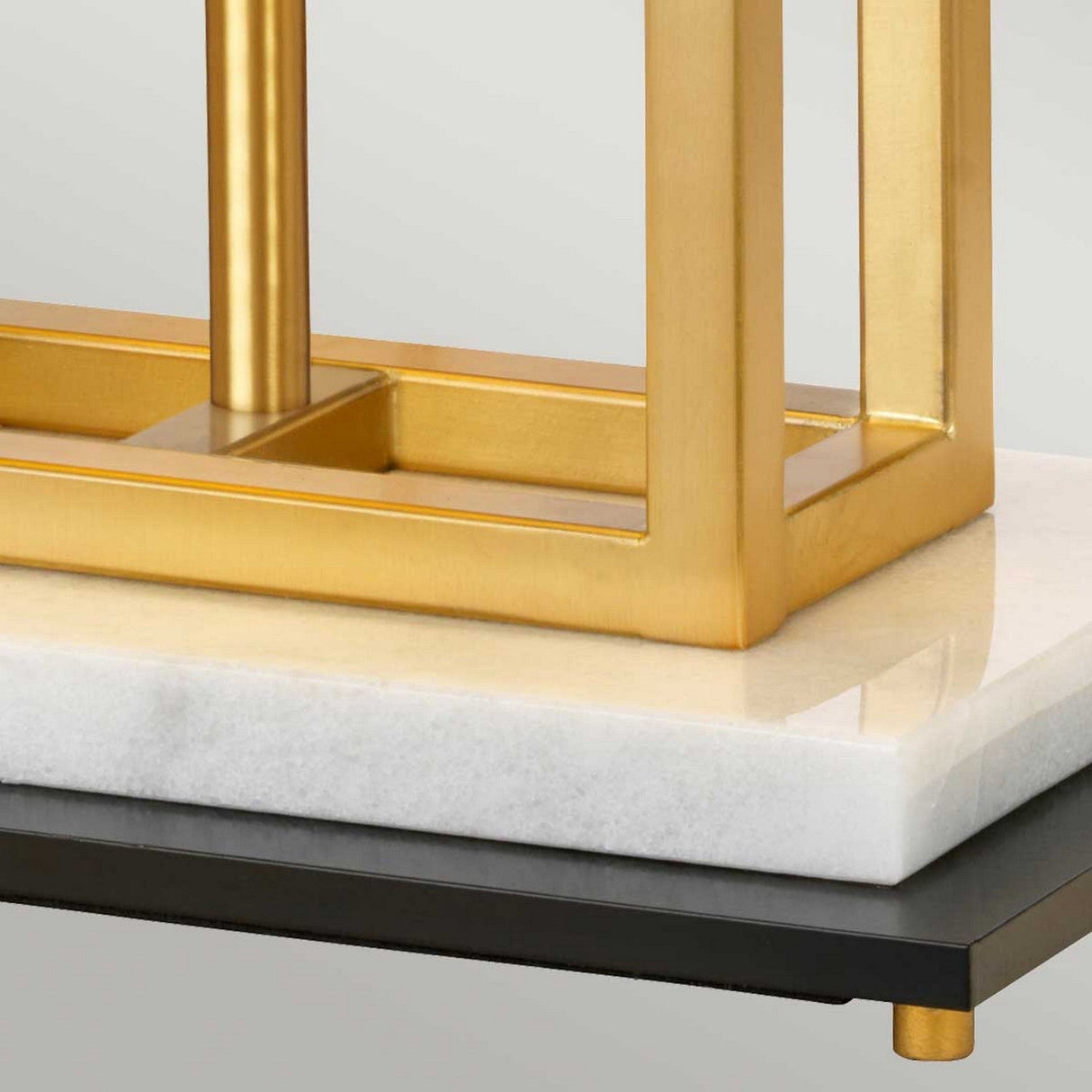A detailed view of the Atlas Table Lamp - Brushed Brass showcases its sleek rectangular frame beautifully set on a white marble base. The marble piece is elegantly positioned on a black surface supported by petite gold feet, adding depth and sophistication to the design.