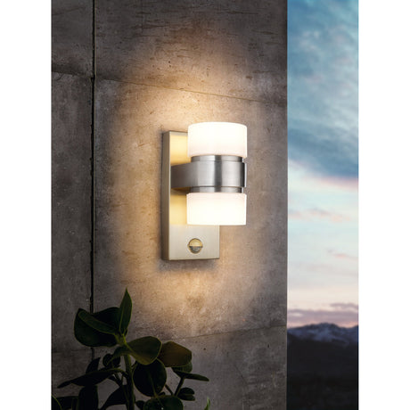 Atollari LED outdoor wall light with stainless steel body, silver central band, and two white cylindrical shades casting warm light up and down a stone exterior wall.