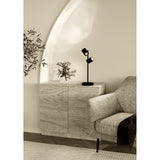 Auro LED Table Lamp in black steel with two adjustable cylindrical spot heads on a slim stem and round base, shown on a wooden sideboard beside an armchair.
