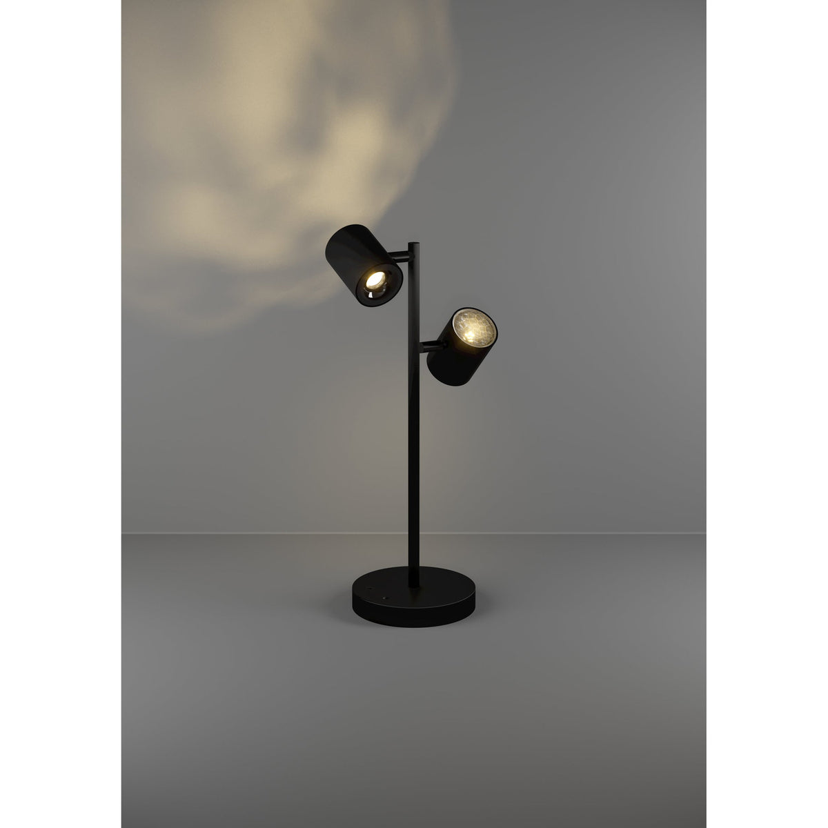 Auro LED Table Lamp in black steel with two adjustable cylindrical spot heads on a slim stem and round disc base, shown lit against a plain grey background.