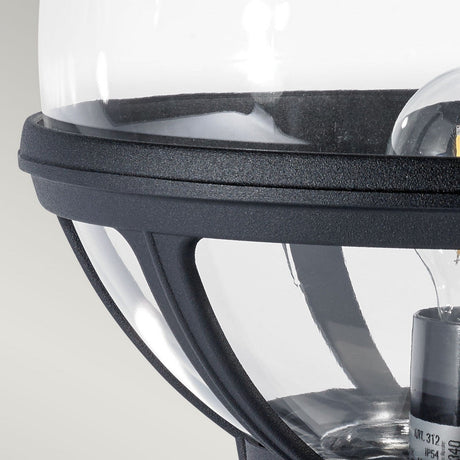 Close-up of the Bologna 1 Light Pedestal - Black, a modern outdoor lighting fixture featuring a clear glass cover and black metal frame set against a light gray background. The design highlights a cylindrical bulb housing within its sleek structure.