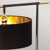 Close-up of the Balance Floor Lamp - Black/Polished Brass, showcasing a black cylindrical shade with a golden interior. The contemporary metal frame includes a horizontal bar on top, reminiscent of its signature style. The lamp emits a warm glow, accentuating the contrasting textures and colors of black and polished brass.