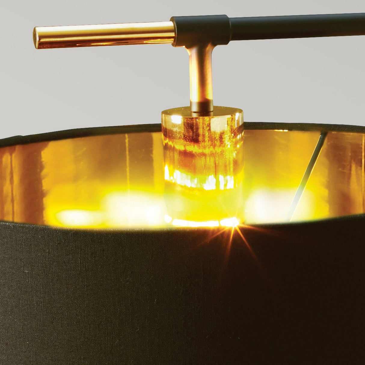 A detailed view of the Balance Table Lamp - Black/Polished Brass exhibits its contemporary design, with a black lampshade supported by cylindrical glass and accented with polished brass details. When illuminated, it emits a warm glow that emphasizes the metallic interior of the shade.