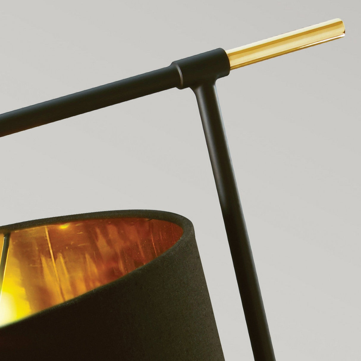 A close-up of the Balance Table Lamp - Black/Polished Brass showcases its modern design with a cylindrical black shade and golden interior. The sleek black stand is accentuated by a polished brass horizontal rod, set against a light gray background.