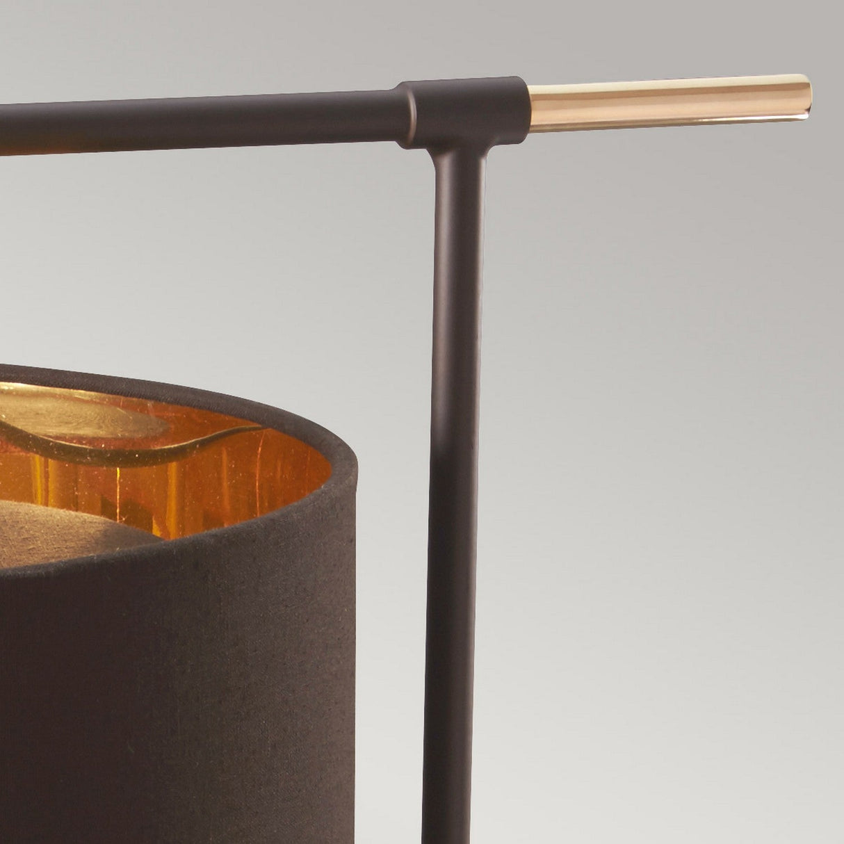 Close-up of the Balance Table Lamp - Brown/Polished Brass showcasing a modern design with a black cylindrical shade and an elegant, polished brass horizontal rod. The interior's reflective golden finish adds sophistication, while the energy-efficient lighting casts a warm glow against its smooth, light gray background.