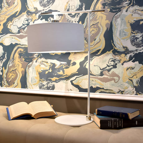 The Balance 1 Light Table Lamp in white and polished nickel sits elegantly on a table, featuring its sleek design with a cylindrical shade. An open book lies nearby, accompanied by two closed books stacked beside it. The background showcases a colorful, swirling marble-patterned wallpaper that enhances the ambiance with its modern flair.
