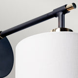 The Balance 1 Light Wall Light in Black and Polished Nickel features a cylindrical white fabric lampshade, paired with a sleek black and polished nickel arm. It connects gracefully to a round black mount on a plain light gray wall, offering a stylish and energy-efficient lighting solution.