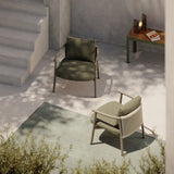 Two Balm aluminium garden lounge chairs with curved arm frames, one upholstered in olive green, the other in beige rope, set on a patio with a rug.