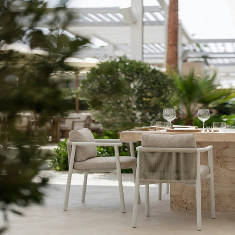 Balm rope garden dining armchair with white aluminium frame, braided rope backrest, and beige cushions, shown at an outdoor dining table amid garden greenery.