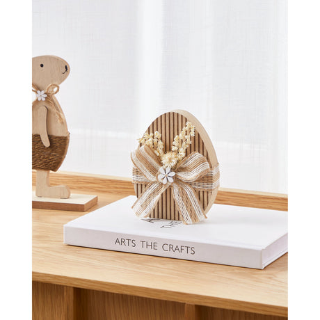 Balmaha Decorative Egg in brown and beige with ribbed stripes, wrapped in a jute and white ribbon bow with dried flowers, displayed on a book beside a wooden rabbit figurine.