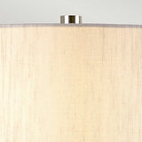 A close-up of the Barbican 1 Light Table Lamp showcases a graphite and silver shade with a smooth texture, resting on its ceramic base. The top is accented by a metallic finial, and the light casts a soft, warm glow on the material. This lamp elegantly combines style and function, adding charm to any room.