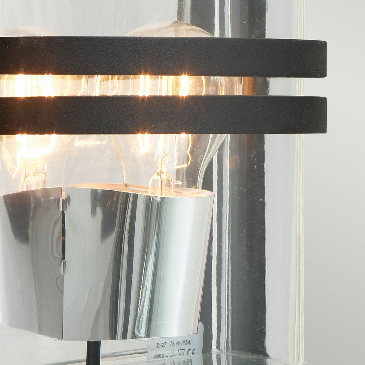 Close-up of the Basel Outdoor 1 Light Wall Lantern, featuring exposed light bulbs inside a clear cylindrical glass casing. This sleek fixture comes in a black finish, accentuated by two parallel black horizontal bands, ideal for bringing contemporary style to any exterior space.