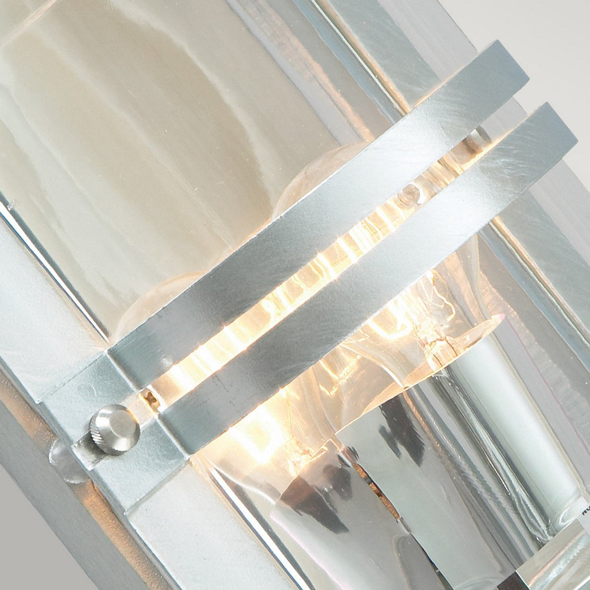 A detailed view of the Basel Outdoor 1 Light Wall Lantern reveals its contemporary metallic structure featuring an exposed bulb, characterized by clean lines and a sleek silver finish. Warm light emanates through its clear glass, highlighting the lantern's modern industrial design.