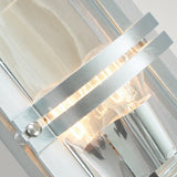 A detailed view of the Basel Outdoor 1 Light Wall Lantern reveals its contemporary metallic structure featuring an exposed bulb, characterized by clean lines and a sleek silver finish. Warm light emanates through its clear glass, highlighting the lantern's modern industrial design.