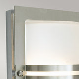 Close-up of a modern wall sconce similar to the Basel Outdoor 1 Light Wall Lantern, featuring a brushed stainless steel frame and frosted glass panel. This design includes two horizontal metal bands and casts a gentle, diffused light against a simple light gray background.