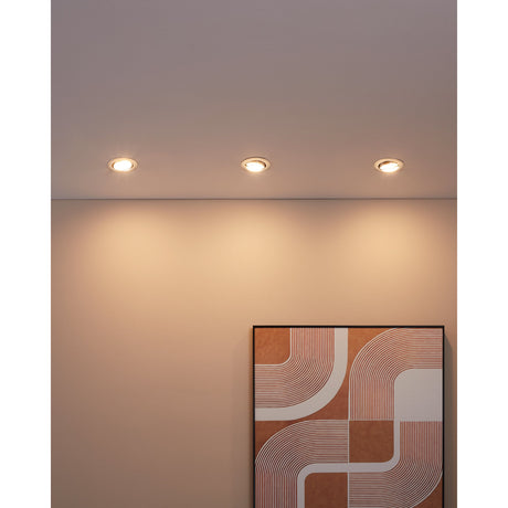 Three round recessed downlights in chrome finish fitted into a ceiling, casting warm light down a wall above a framed abstract print in a terracotta and white pattern.