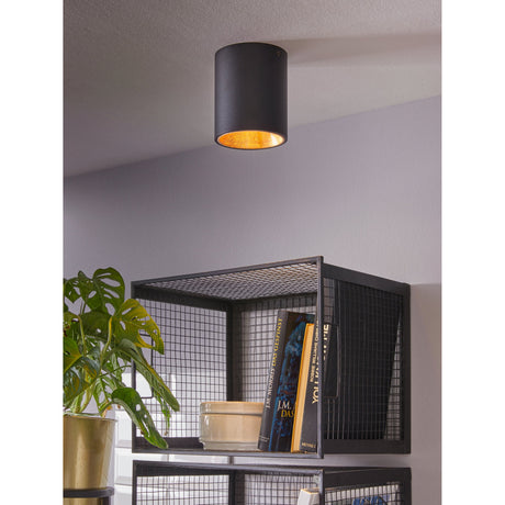 Basmorne LED Flush Ceiling Light in matt black cylindrical aluminium with gold-leaf effect interior, mounted above a wire-mesh shelving unit in a living room setting.