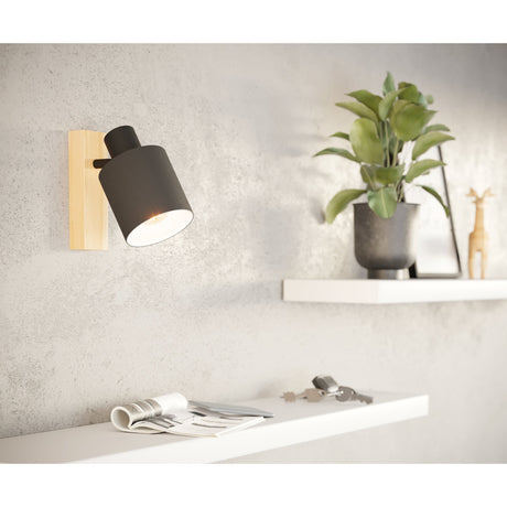 Batallas Wall Spotlight in brown and black, with a pale wood wall block and adjustable black fabric shade, shown lit beside a shelf with a plant and keys.