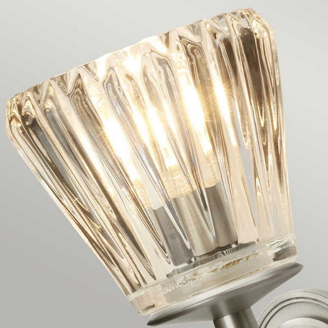 A close-up of the Agatha 2 Light Wall Light - Brushed Nickel reveals a lit, ribbed glass sconce emitting a warm glow. The brushed nickel finish on the metal fixture gives it a modern touch, and its IP44 rating ensures it is durable in damp environments.