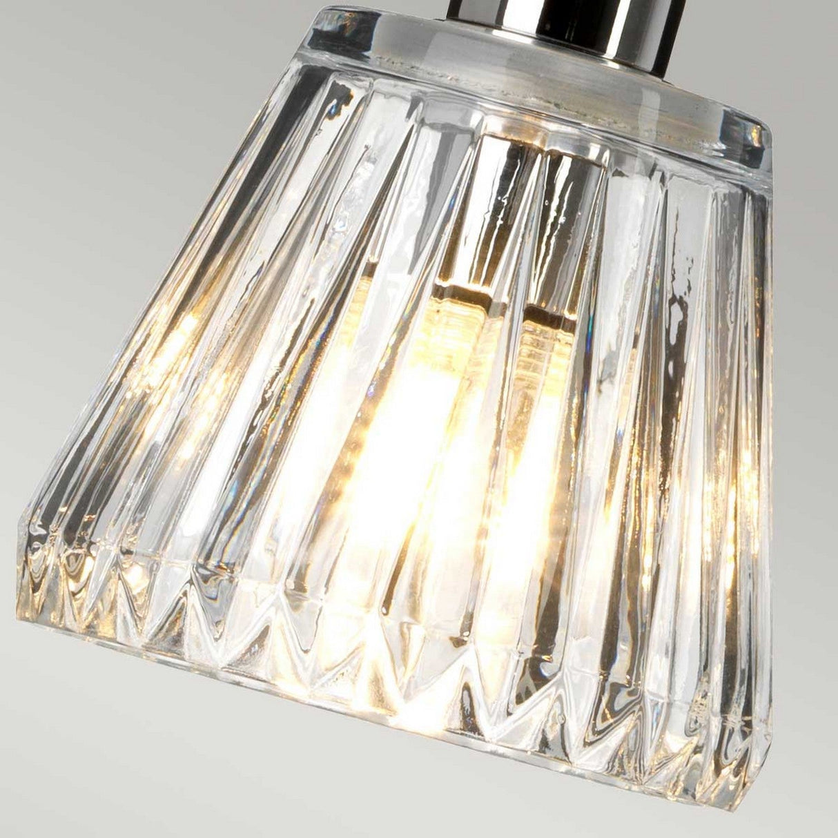 A close-up of the Agatha 1 Light Pendant in polished chrome reveals a modern clear glass design with geometric features, beautifully illuminated from within. The bulb emits a warm glow that enhances the prismatic texture of the glass and is complemented by sleek chrome accents.