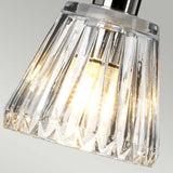 A close-up of the Agatha 1 Light Pendant in polished chrome reveals a modern clear glass design with geometric features, beautifully illuminated from within. The bulb emits a warm glow that enhances the prismatic texture of the glass and is complemented by sleek chrome accents.
