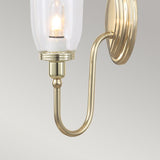 The Blake Wall Light - Polished Brass showcases a polished brass sconce with a gracefully curved arm, which holds a clear glass shade encasing an LED G9 lamp. This sophisticated light fixture is mounted on a round brass base and set against a light gray background.