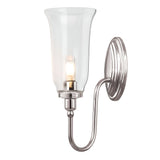 The Blake Wall Light - Polished Nickel showcases a sleek polished nickel base and a gracefully curved arm supporting a clear, flared glass shade. Inside, it houses a light bulb that emits a warm glow, enhancing the modern aesthetic of this wall-mounted sconce.