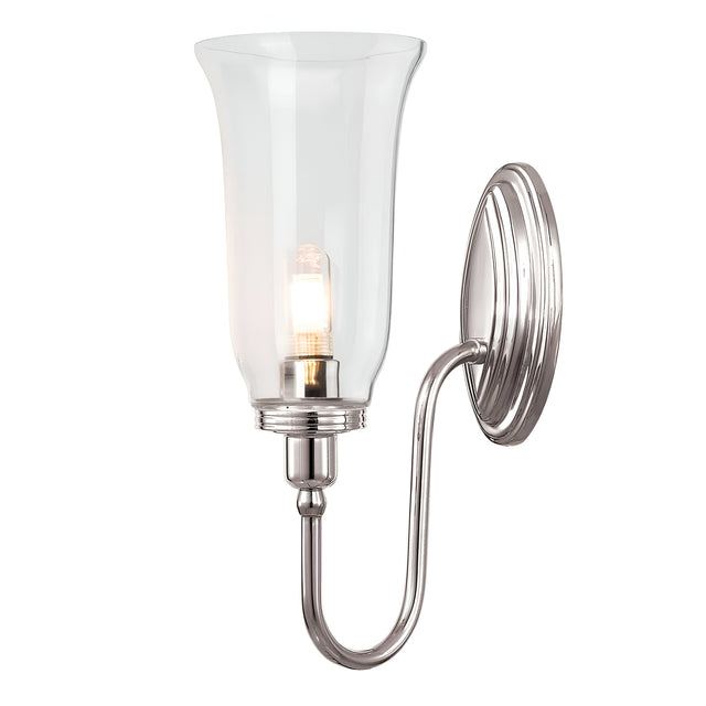 The Blake Wall Light - Polished Nickel showcases a sleek polished nickel base and a gracefully curved arm supporting a clear, flared glass shade. Inside, it houses a light bulb that emits a warm glow, enhancing the modern aesthetic of this wall-mounted sconce.