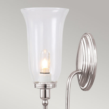 The Blake Wall Light - Polished Nickel boasts a polished nickel base paired with a frosted glass shade. It softly diffuses the bulb's light, casting a gentle glow against the light gray backdrop, making it an elegant addition to any space.