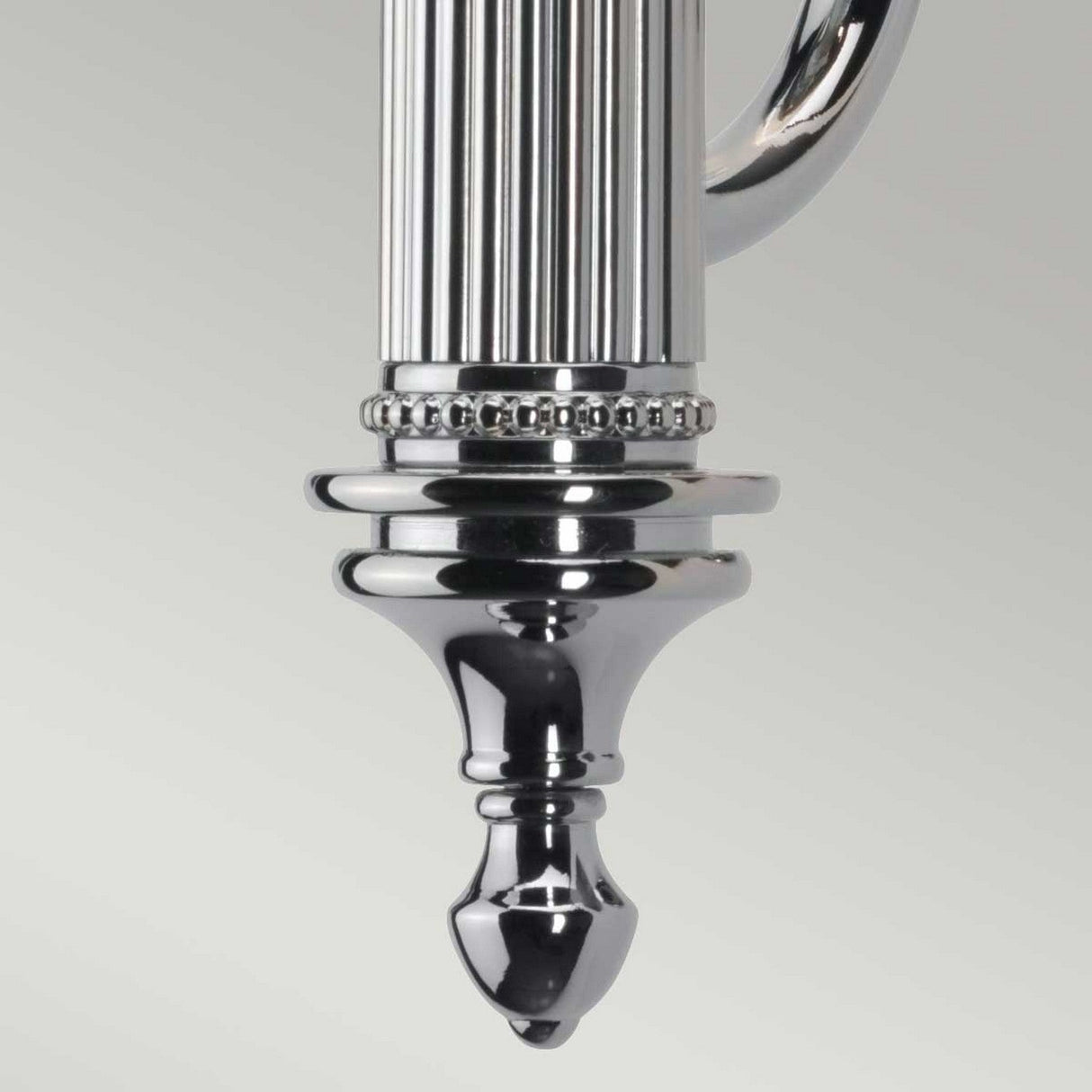 Close-up of the Carroll-2 Wall Light in polished chrome, featuring intricate grooved and beaded patterns that culminate in a pointed finial. The reflective surface gleams against a light gray background, highlighting its role as a contemporary lighting fixture.