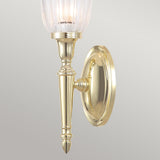 Introducing the Dryden-1 Wall Light in polished brass, characterized by its oval base and sleek vertical design. It boasts a ribbed glass lampshade at the top, using an LED G9 lamp to enhance its classic charm against a neutral gray background.