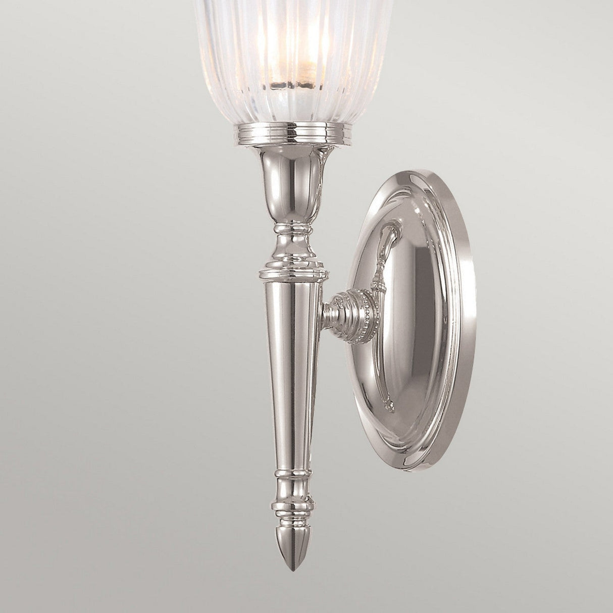 Introducing the Dryden-1 Wall Light - Polished Nickel: This stylish wall sconce showcases a sleek design with a ribbed glass shade set against a plain backdrop. Its elegant, elongated form is enhanced with decorative detailing on the arm and base of the fixture. Compatible with an LED G9 lamp for energy-efficient lighting, it is ideal for modern interiors.