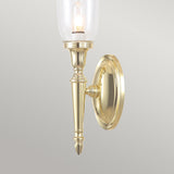 The Dryden-2 Wall Light in Polished Brass showcases a tall, clear glass shade with a visible bulb, elegantly designed for traditional interiors. It features intricate detailing and a polished finish, all mounted on a round brass base.
