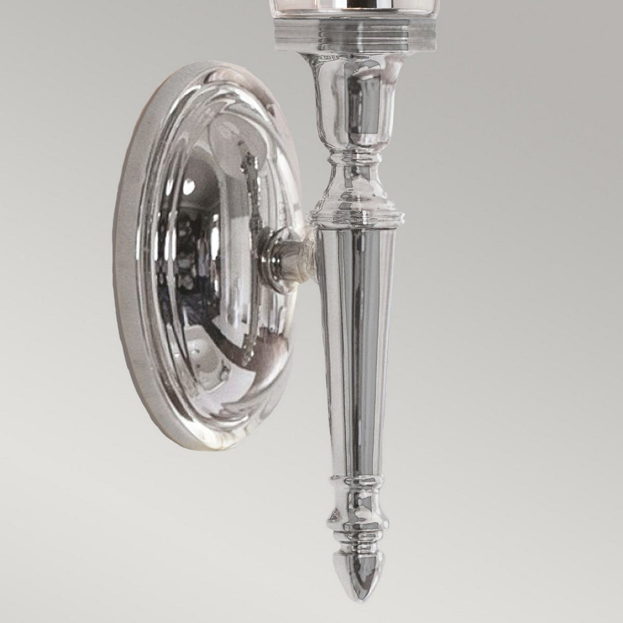 The Dryden-2 Wall Light - Polished Chrome is an elegant sconce with a traditional elongated shape and intricate detailing. It includes compatibility with LED G9 lamps, and its polished chrome surface is mounted on a circular base against a light gray backdrop.