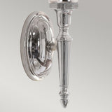 The Dryden-2 Wall Light - Polished Chrome is an elegant sconce with a traditional elongated shape and intricate detailing. It includes compatibility with LED G9 lamps, and its polished chrome surface is mounted on a circular base against a light gray backdrop.