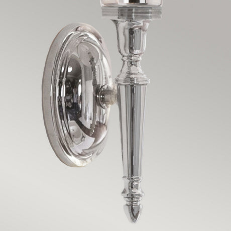 The Dryden-2 Wall Light - Polished Chrome is an elegant sconce with a traditional elongated shape and intricate detailing. It includes compatibility with LED G9 lamps, and its polished chrome surface is mounted on a circular base against a light gray backdrop.