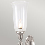 The Dryden-2 Wall Light in Polished Nickel is a stylish wall-mounted fixture featuring a tall, translucent glass shade. It is illuminated by LED G9 lamps that cast a warm glow against the plain, light-colored wall.