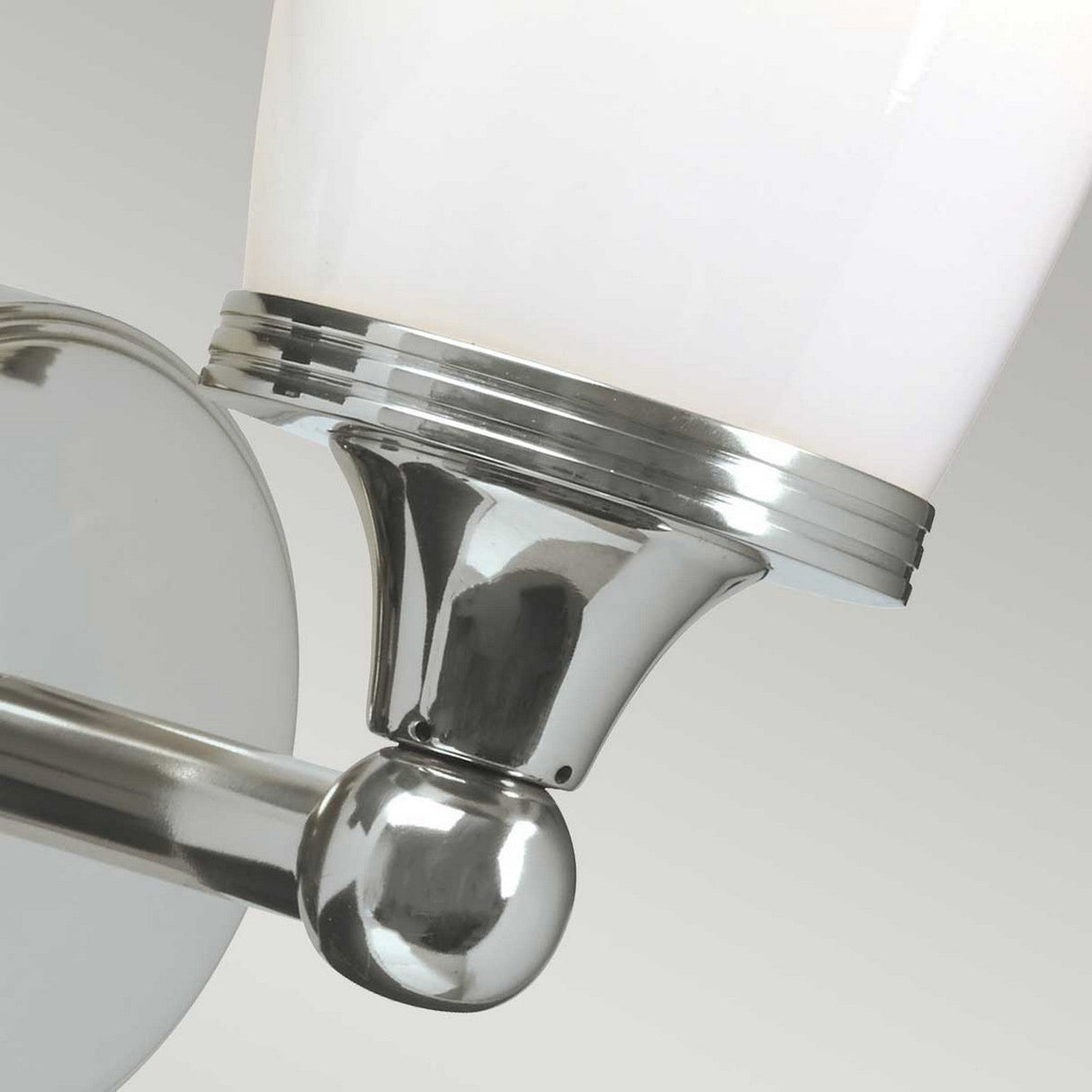 A close-up of the Eliot 1 Light Wall Light in polished chrome showcases its sleek, modern design. The fixture includes an upward-facing white frosted glass shade, held by a shiny metallic base and attached to a circular wall mount. Perfect for bathroom lighting, it elegantly complements the soft gray gradient backdrop.