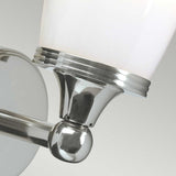 A close-up of the Eliot 1 Light Wall Light in polished chrome showcases its sleek, modern design. The fixture includes an upward-facing white frosted glass shade, held by a shiny metallic base and attached to a circular wall mount. Perfect for bathroom lighting, it elegantly complements the soft gray gradient backdrop.