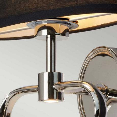 A close-up of the Falmouth Wall Light - Polished Chrome/Black Shade showcases its elegant, curved design and circular lamp with a black shade, which casts a warm glow. The sleek, reflective chrome finish contrasts beautifully against the plain off-white background.