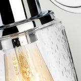 Close-up of the Morvah 1 Light Outdoor Wall Light - Polished Chrome. The clear glass shade boasts water droplet designs, making it an ideal choice for outdoor lighting. Inside, a visible glowing bulb highlights an intricate filament.