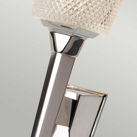Presenting a close-up of the Verity 1 Light Wall Light in polished chrome, featuring a textured square glass shade. This energy-efficient wall-mounted lamp angles slightly, casting captivating reflections on a smooth, light-colored background, making it ideal for modern interiors.