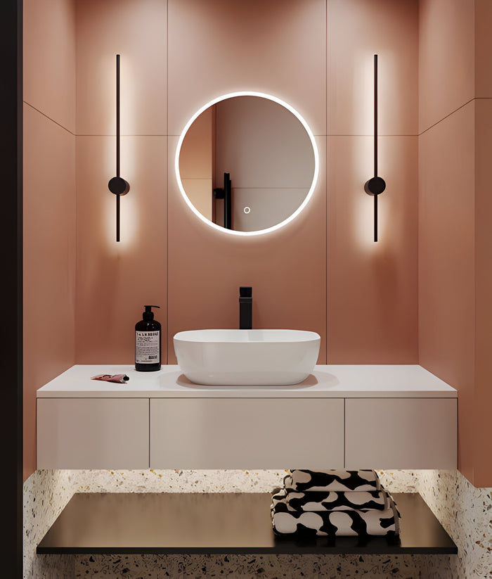 A modern bathroom with peach walls features a round, backlit mirror above a white countertop and oval sink. Two vertical black lights flank the mirror. A bottle of soap and folded towels with a black-and-white pattern are on the counter.