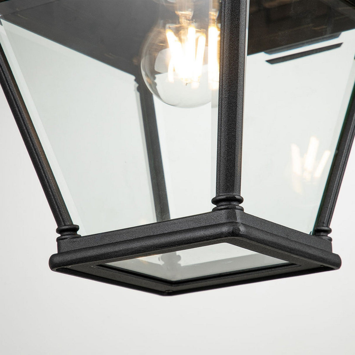 Close-up of the Bayview 1 Light Outdoor Large Wall Lantern in black, showcasing its angular, geometric design with a clear glass enclosure encasing a visible incandescent bulb. The fixture emits a warm glow, ideal for enhancing outdoor spaces.