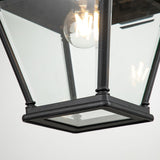 Close-up of the Bayview 1 Light Outdoor Large Wall Lantern in black, showcasing its angular, geometric design with a clear glass enclosure encasing a visible incandescent bulb. The fixture emits a warm glow, ideal for enhancing outdoor spaces.