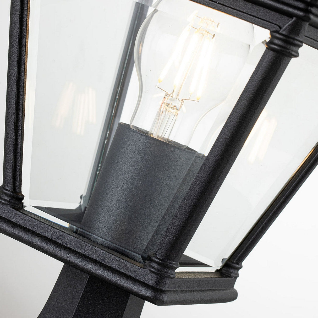 Close-up of the black Bayview 1 Light Medium Pedestal Lantern, featuring clear glass panels that display a glowing filament light bulb inside. This outdoor lighting fixture merges sleek, modern lines with touches of classical Georgian design, making it an elegant addition to any exterior space.