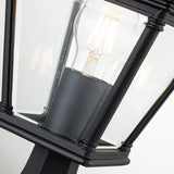 Close-up of the black Bayview 1 Light Medium Pedestal Lantern, featuring clear glass panels that display a glowing filament light bulb inside. This outdoor lighting fixture merges sleek, modern lines with touches of classical Georgian design, making it an elegant addition to any exterior space.