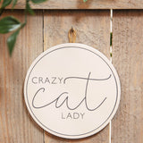 Round cream MDF wall plaque reading "Crazy cat Lady" in dark grey lettering with a fine ring border, hung by a jute cord on a wooden fence panel.