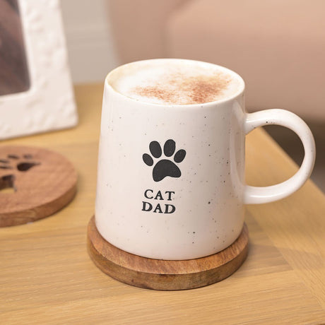 White speckled stoneware Cat Dad mug with black paw print and loop handle, filled with frothy coffee, shown on a wooden coaster beside a paw-print coaster.
