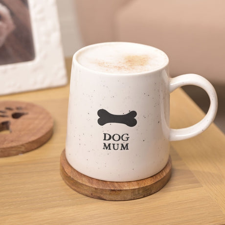 White speckled stoneware Dog Mum mug with black bone print and text, filled with frothy coffee, shown on a wooden coaster beside a paw-print trivet.